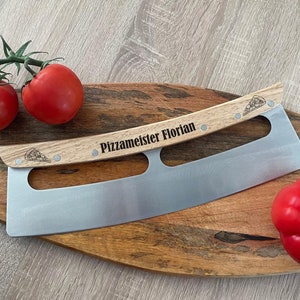 Może przedstawiać: N&oacute;ż do pizzy ze stali nierdzewnej z zakrzywionym ostrzem i jasnym drewnianym uchwytem. Uchwyt jest wygrawerowany napisem "Pizzameister Florian" i posiada małe ikony kawałk&oacute;w pizzy na obu końcach, przymocowane za pomocą czterech metalowych nit&oacute;w.