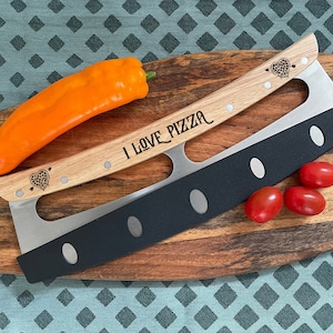 Może przedstawiać: Krajacz do pizzy z drewnianą rączką z wygrawerowanym napisem I LOVE PIZZA oraz dwiema ikonami serc ze strzałami. Narzędzie posiada długie, zakrzywione ostrze ze stali nierdzewnej z matowym czarnym wykończeniem w dolnej części oraz srebrnymi nitami.