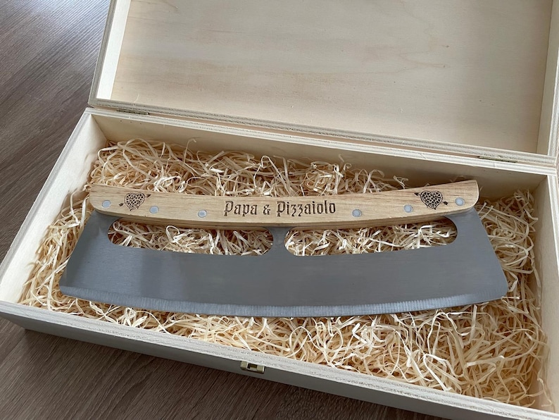 Może przedstawiać: Zakrzywiony metalowy n&oacute;ż do pizzy z jasnym drewnianym uchwytem, na kt&oacute;rym wygrawerowano napis Papa & Pizzaiolo oraz dwa serca. N&oacute;ż znajduje się w prostokątnym drewnianym pudełku wypełnionym jasnymi wi&oacute;rkami drzewnymi.