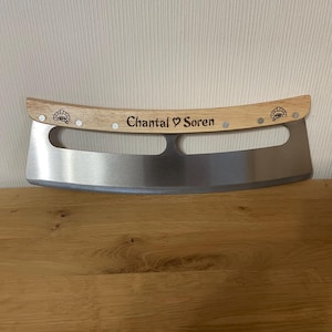 Może przedstawiać: Zakrzywiony n&oacute;ż do pizzy ze stali nierdzewnej z jasnym drewnianym uchwytem. Na uchwycie znajduje się wygrawerowany napis "Chantal ♡ Soren", umieszczony pomiędzy dwiema małymi ikonami pieca do pizzy po lewej i prawej stronie drewna.