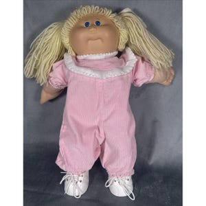 May include: A vintage doll with a soft, vinyl face, blue eyes, and blonde yarn hair styled in pigtails. The doll wears a pink corduroy jumpsuit with a white ruffled collar and white lace-up shoes. A classic toy.