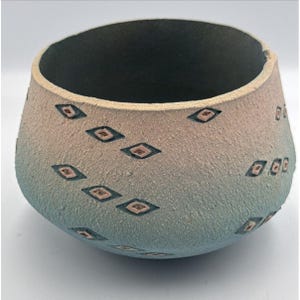 Puede incluir: Un cuenco de cerámica hecho a mano con una superficie texturizada y una combinación de colores degradados. El cuenco pasa de un beige claro en la parte superior a un azul verdoso en la base. Presenta un patrón de diamantes repetido con un contorno azul oscuro y un centro marrón rojizo.