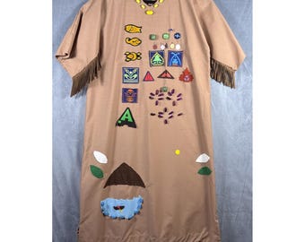 Könnte beinhalten: Ein beigefarbenes Tunika-Kleid mit kurzen Ärmeln und Fransenbesatz. Das Kleid ist mit bunten Aufnähern verziert, darunter Fische, geometrische Formen und ein "A"-Aufnäher. Ein dekorativer Ausschnitt und ein hausförmiges Design sind ebenfalls zu sehen.