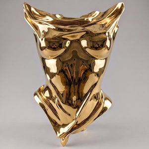 Puede incluir: Una escultura de color dorado pulido que representa un torso humano con una superficie altamente reflectante similar a un espejo. La forma presenta contornos fluidos y drapeados que imitan pliegues de tela, creando un aspecto metálico liso.