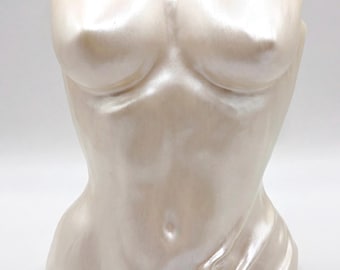 May include: A pearlescent off-white torso sculpture featuring a smooth, contoured human form with a subtle draped fabric effect at the lower side. The surface has a soft, reflective sheen, highlighting the anatomical curves of the chest and abdomen.
