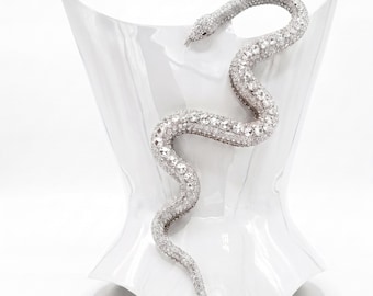 May include: A silver-toned snake-shaped accessory covered in clear, sparkling rhinestones. The snake is depicted in a winding, S-shaped posture with a pointed tail and a detailed head, featuring a reflective surface that catches light across its body.