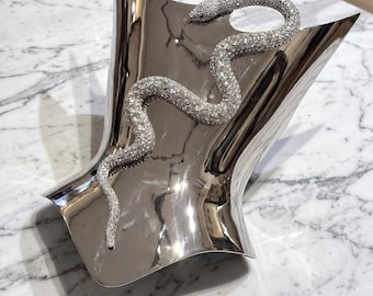 May include: A polished silver-toned metallic display stand with a curved, concave shape. A long, winding snake figurine covered in small, sparkling clear crystals is attached to the front surface, curving from the top right to the bottom left.