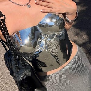 May include: A strapless, reflective silver metallic corset top featuring a central, intricate silver-toned ornamental embellishment. The garment is paired with a black quilted shoulder bag with a chain strap and a textured silver-toned skirt.