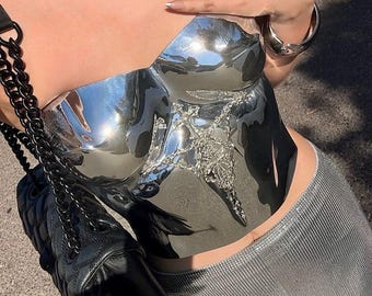 May include: A strapless, reflective silver metallic corset top featuring a central, intricate silver-toned ornamental embellishment. The garment is paired with a black quilted shoulder bag with a chain strap and a textured silver-toned skirt.