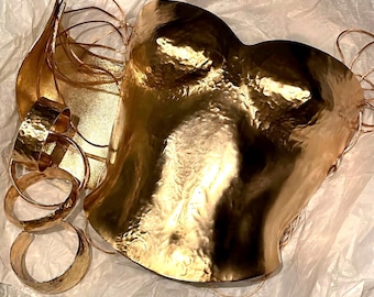 Op de afbeelding: Een goudkleurige metalen korsettop met een gehamerd, getextureerd oppervlak en een hartvormige halslijn, vergezeld van drie bijpassende gehamerde metalen armbanden en een apart gebogen metalen stuk, allemaal met een reflecterende afwerking.