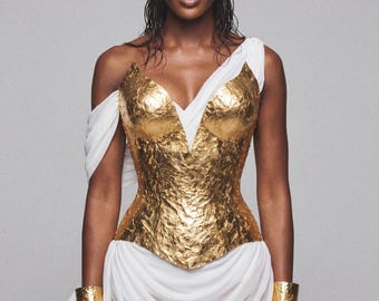 Peut inclure: Une femme porte un bustier doré texturé avec un décolleté en V et un tissu blanc drapé. Des bracelets de bras dorés complètent l'ensemble. L'ensemble évoque l'esthétique grecque antique.
