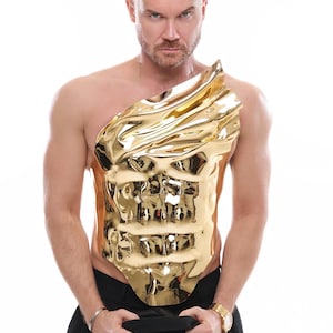 May include: A gold-colored, sculpted chest piece with an abstract design. The piece is worn over the torso. The person in the image is holding a black garment and wearing a gold watch and silver bracelet.