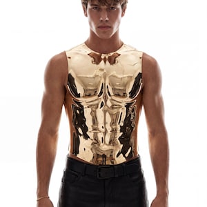 May include: A sleeveless, metallic gold chest plate with a highly reflective, polished finish. The garment features a molded, anatomical design that contours to the torso, extending from the collarbone down to the waist, paired with black pants.