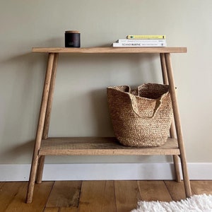 May include: A light brown wooden console table with a lower shelf and angled legs. A woven basket, a black candle, and two books are on the shelves. The books are titled "SCANDI RUSTIC" and "JAPANESE STYLE".