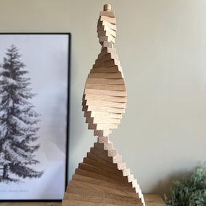 May include: A wooden sculpture with a spiral design. The sculpture is made of light-colored wood and has a triangular base that tapers upwards to a rounded top. The design is reminiscent of a Christmas tree. The sculpture is displayed on a wooden surface.