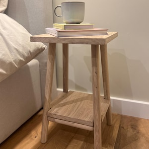 May include: A small, light-colored wooden side table with a square top and a lower shelf. A light green mug and two books are on top. The table has four legs and is placed next to a bed.