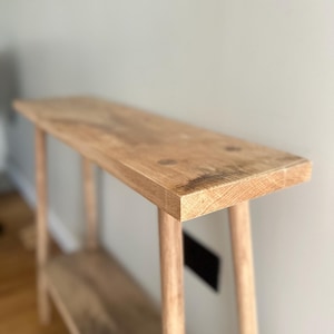 K&ouml;nnte beinhalten: Ein helles Holzkonsolentisch mit rechteckiger Oberseite und einem unteren Regal. Der Tisch hat vier schlanke, abgerundete Beine. Die Holzmaserung ist auf der Oberseite und dem Regal sichtbar. Der Tisch ist etwa 122 cm lang.