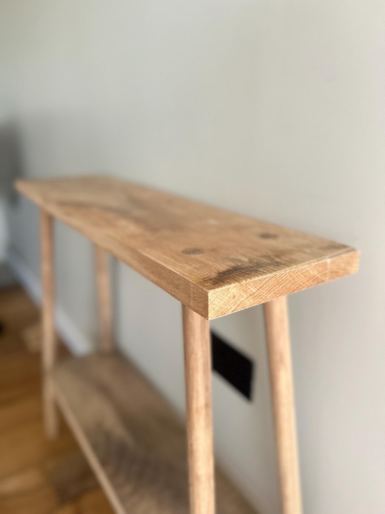 K&ouml;nnte beinhalten: Ein helles Holzkonsolentisch mit rechteckiger Oberseite und einem unteren Regal. Der Tisch hat vier schlanke, abgerundete Beine. Die Holzmaserung ist auf der Oberseite und dem Regal sichtbar. Der Tisch ist etwa 122 cm lang.