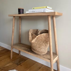 K&ouml;nnte beinhalten: Ein helles Holzkonsolentisch mit einem unteren Regal und schr&auml;gen Beinen. Auf dem unteren Regal steht ein geflochtener Korb. B&uuml;cher und eine Kerze befinden sich auf dem oberen Regal. Das Wort "SCANDIRUSTIC" ist auf einem der B&uuml;cher zu sehen.