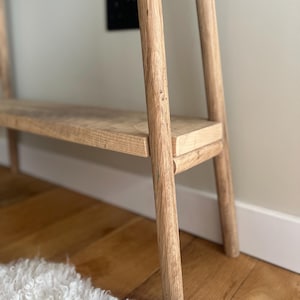 K&ouml;nnte beinhalten: Eine helle Holzkonsolentisch mit einem Regal. Der Tisch hat ein minimalistisches Design mit vier abgerundeten Beinen und einem rechteckigen Regal. Die Holzmaserung ist sichtbar. Der Tisch steht auf einem Hartholzboden, mit einem wei&szlig;en Teppich im Vordergrund.