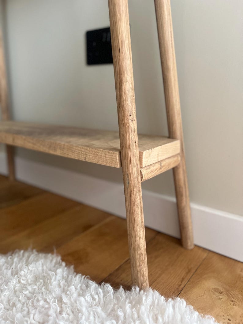 K&ouml;nnte beinhalten: Eine helle Holzkonsolentisch mit einem Regal. Der Tisch hat ein minimalistisches Design mit vier abgerundeten Beinen und einem rechteckigen Regal. Die Holzmaserung ist sichtbar. Der Tisch steht auf einem Hartholzboden, mit einem wei&szlig;en Teppich im Vordergrund.