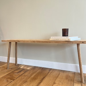 May include: A wooden bench with four legs, a light brown finish, and a natural wood grain. The bench is sitting on a wooden floor. Two books and a glass candle holder are on top of the bench.