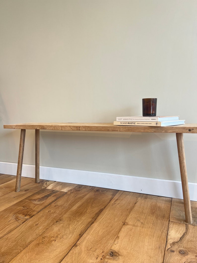 May include: A wooden bench with four legs, a light brown finish, and a natural wood grain. The bench is sitting on a wooden floor. Two books and a glass candle holder are on top of the bench.