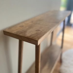 K&ouml;nnte beinhalten: Ein hellbrauner Konsolentisch aus Holz mit einer langen, rechteckigen Oberseite und einem unteren Regal. Der Tisch hat vier runde Beine und ist aus Naturholz gefertigt. Der Tisch ist ca. 152 cm lang, 30 cm breit und 76 cm hoch.