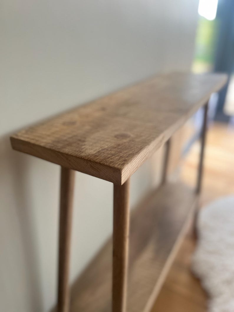 K&ouml;nnte beinhalten: Ein hellbrauner Konsolentisch aus Holz mit einer langen, rechteckigen Oberseite und einem unteren Regal. Der Tisch hat vier runde Beine und ist aus Naturholz gefertigt. Der Tisch ist ca. 152 cm lang, 30 cm breit und 76 cm hoch.