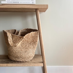 K&ouml;nnte beinhalten: Eine helle Holzkonsolentisch mit einem geflochtenen Jute-Korb auf dem unteren Regal. Drei B&uuml;cher sind auf dem oberen Regal gestapelt. Die B&uuml;cher tragen die Titel "SCANDIRUSTIC", "JAPANDI STYLE" und "THE SCANDINAVIAN GUIDE TO HAPPINESS".