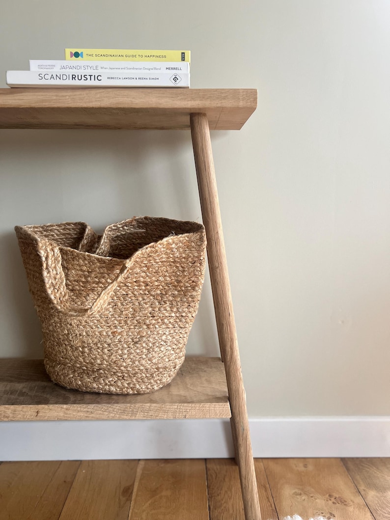 K&ouml;nnte beinhalten: Eine helle Holzkonsolentisch mit einem geflochtenen Jute-Korb auf dem unteren Regal. Drei B&uuml;cher sind auf dem oberen Regal gestapelt. Die B&uuml;cher tragen die Titel "SCANDIRUSTIC", "JAPANDI STYLE" und "THE SCANDINAVIAN GUIDE TO HAPPINESS".