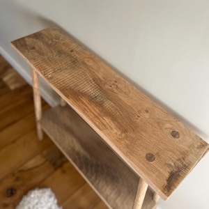 K&ouml;nnte beinhalten: Handgefertigte Holztischkonsole mit nat&uuml;rlichem Finish. Der Tisch hat eine lange, rechteckige Oberseite und ein unteres Regal, das von vier schlanken Beinen getragen wird. Die Holzmaserung ist sichtbar. Der Tisch ist ca. 122 cm lang.