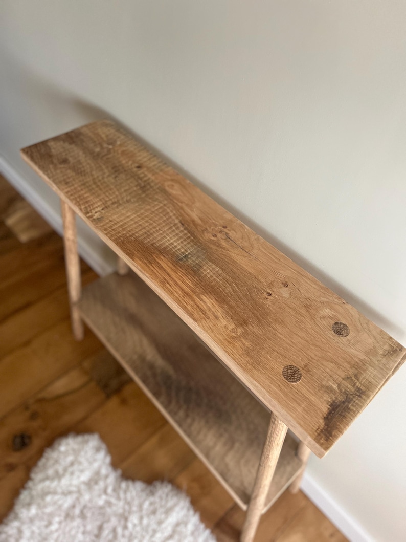 K&ouml;nnte beinhalten: Handgefertigte Holztischkonsole mit nat&uuml;rlichem Finish. Der Tisch hat eine lange, rechteckige Oberseite und ein unteres Regal, das von vier schlanken Beinen getragen wird. Die Holzmaserung ist sichtbar. Der Tisch ist ca. 122 cm lang.