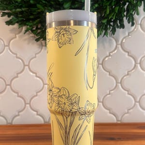 May include: A yellow stainless steel tumbler with a floral design of daffodils. The tumbler has a lid and a straw.