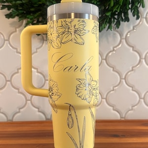 May include: A yellow stainless steel travel mug with a black handle and a straw. The mug features a floral design and the name "Carla" on it.