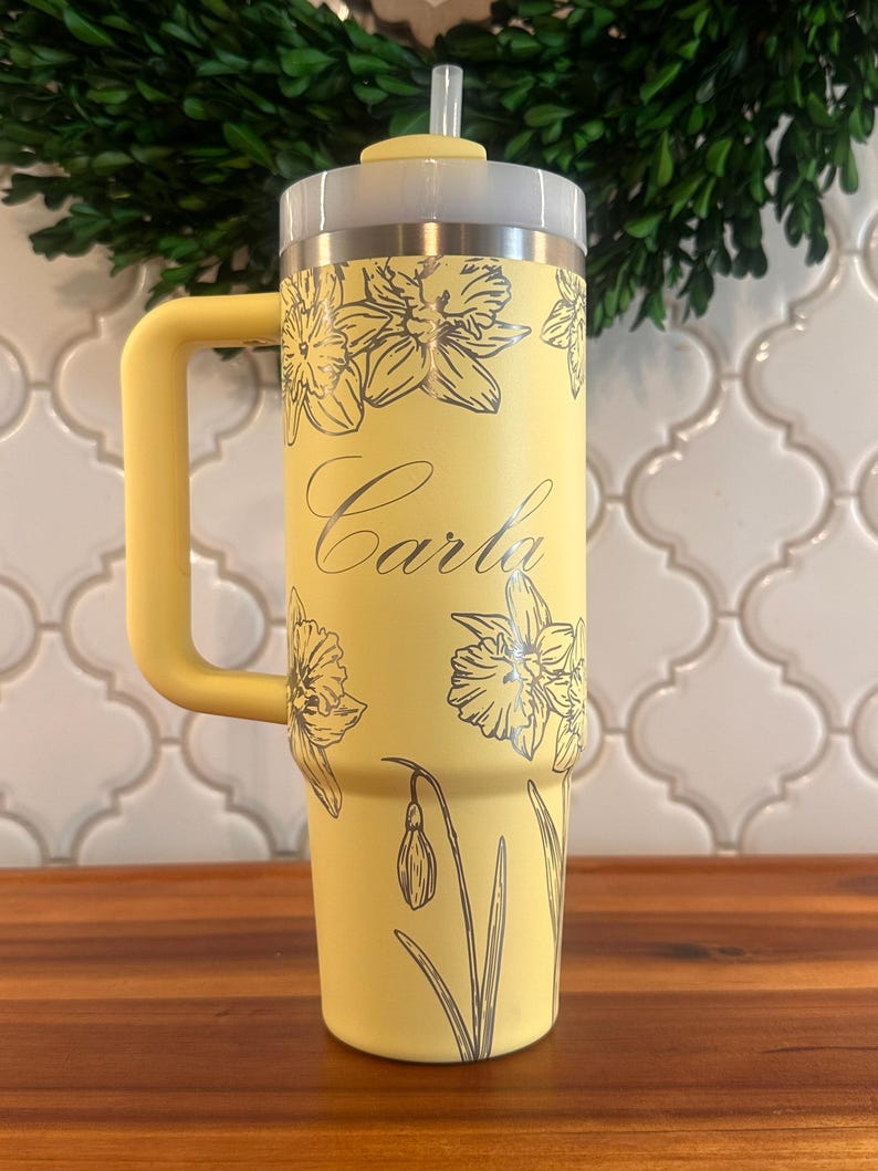 May include: A yellow stainless steel travel mug with a black handle and a straw. The mug features a floral design and the name "Carla" on it.