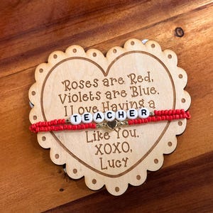 May include: A wooden heart-shaped sign with the text "Roses are Red, Violets are Blue. I love Having a TEACHER Like You. XOXO, Lucy". A red beaded bracelet with gold beads and the word "TEACHER" spelled out in white letters is on top of the sign.