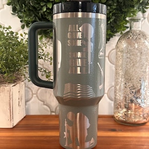 May include: A green stainless steel travel mug with a black lid and a handle. The mug has a design of a soldier kneeling, an American flag, and the text "All gave some, some gave all".