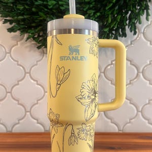 May include: A yellow stainless steel travel mug with a black floral design and a black handle. The mug has a straw lid and the text "STANLEY" is printed on the side.