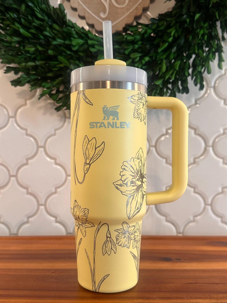 May include: A yellow stainless steel travel mug with a black floral design and a black handle. The mug has a straw lid and the text "STANLEY" is printed on the side.