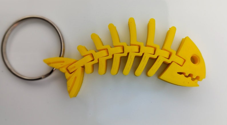 May include: A yellow plastic fish skeleton keyring. The skeleton is detailed and has a realistic shape. The keyring is attached to a silver metal ring.
