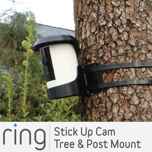 May include: A black Ring Stick Up Cam mounted on a tree using a black strap mount. The camera is white with a black lens and a black cover. The text "Stick Up Cam Tree & Post Mount" is visible below the image.
