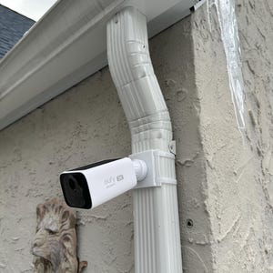 May include: A white and black Eufy 2K security camera mounted on a white downspout. The camera is positioned to monitor an outdoor area. The downspout is attached to a textured beige wall. An ornate lion head is mounted on the wall.