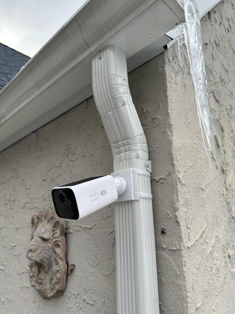 May include: A white and black Eufy 2K security camera mounted on a white downspout. The camera is positioned to monitor an outdoor area. The downspout is attached to a textured beige wall. An ornate lion head is mounted on the wall.