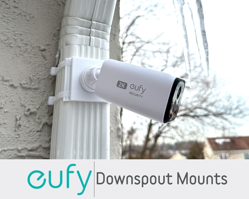 May include: A white security camera mounted on a white downspout mount. The camera has a black lens and the text "2K eufy SECURITY". The image also includes the text "eufy Downspout Mounts".