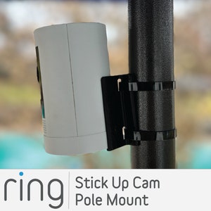 May include: A white Ring Stick Up Cam mounted on a black pole mount. The camera is facing forward and the pole mount is attached to a black pole.
