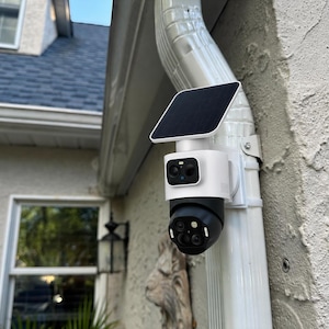 May include: A white and black security camera mounted on a white downspout. The device features a square solar panel on top, a white cylindrical housing, and a black rotating camera lens assembly with multiple sensors and a small blue indicator light.
