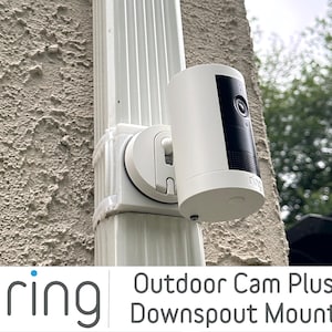 May include: White outdoor security camera with a black front panel, mounted on a white downspout mount. The camera is attached to a white downspout. The image includes the "ring" logo and the words "Outdoor Cam Plus Downspout Mount".