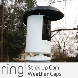 May include: A white Ring Stick Up Cam with a black weather cap. The camera is mounted on a white base and is positioned in front of a blurred background of trees.