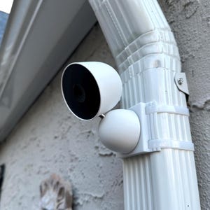May include: A white, spherical security camera with a black circular lens face mounted to a white vertical downspout using a custom bracket and two white zip ties. The camera housing is smooth and rounded, positioned at an angle to monitor.